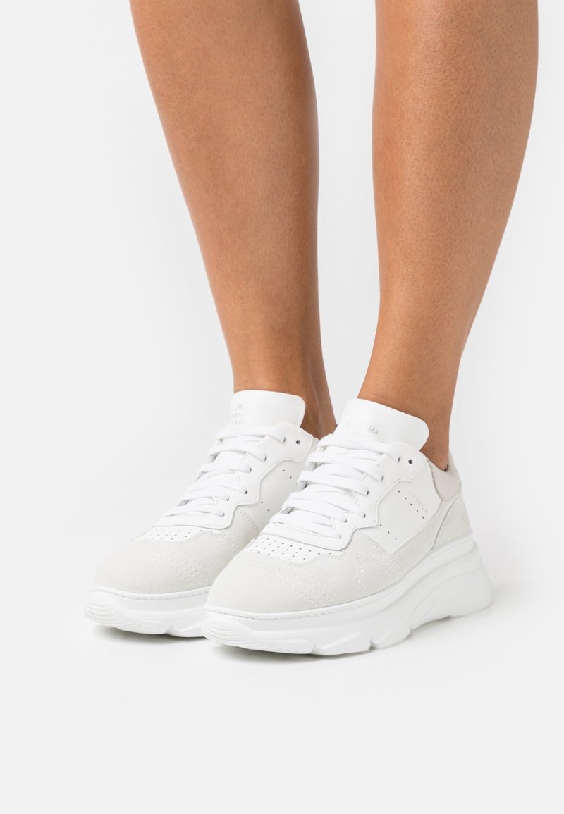 Baskets blanches à lacets avec semelle épaisse portées sur des jambes nues sur un fond blanc uni, vues de face et de profil.