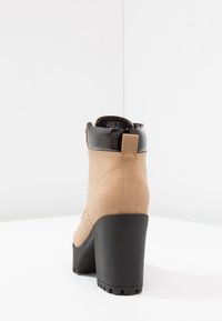 Botines de tobillo color marrón claro con tacón y caña negra brillante. Presentan un tacón bloque grueso y una suela de goma texturizada para mayor agarre.
