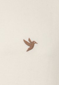 Crème gebreide trui met een klein bruin geborduurd vogellogo aan de linkerkant, met een gladde textuur en subtiele ribbeldetails.