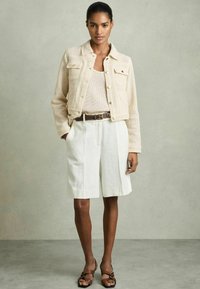 Beige gebreide jas met knoopsluitingen en zakken, gecombineerd met een textuur tanktop en witte op maat gemaakte shorts. Bruine riem en sandaaltjes met bandjes.