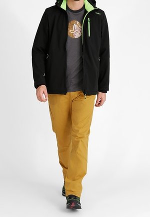 Veste softshell noire avec accents verts, capuche et fermetures éclair. T-shirt gris en dessous, associé à un pantalon jaune et des chaussures noires.
