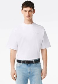 Camiseta de algodón blanca con cuello redondo y mangas cortas. Presenta un sutil texto bordado en gris en la parte frontal. Se lleva con denim claro y un cinturón negro.