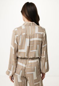 Blusa decorata con un design geometrico in tonalità beige e marrone. Presenta maniche lunghe, colletto e un dettaglio in vita elasticizzato.