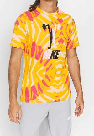 Persoon draagt een geel-roze tie-dye Nike T-shirt met een zwart-witte gewichtheffen afbeelding, gecombineerd met lichtgrijze Nike short.
