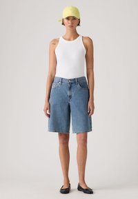 Γυναίκα που φοράει κίτρινο καπέλο, λευκό αμάνικο τοπ, μπλέ τζιν σορτς μέχρι το γόνατο και μαύρα μπαλαρίνια, στέκεται μπροστά σε φωτεινό φόντο.