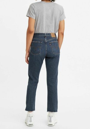 Persoon draagt high-waisted blauwe spijkerbroek en een grijs t-shirt, staat met rug naar voren, witte sneakers zichtbaar.