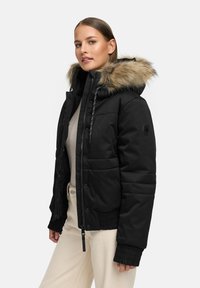Jachetă puffer neagră cu glugă din blană artificială, model matlasat, fermoar frontal și manșete ribbate. Asortată cu un top și pantaloni de culoare deschisă.