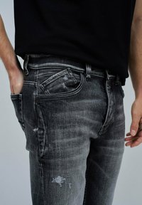 Jeans de mezclilla negra texturizada con un patrón desteñido y desgastado, que presentan un bolsillo lateral y un diseño ajustado.