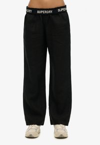 Pantalons larges en lin noir avec une taille élastique arborant le logo "SUPERDRY" en blanc et bleu. Tissu texturé et coupe décontractée.