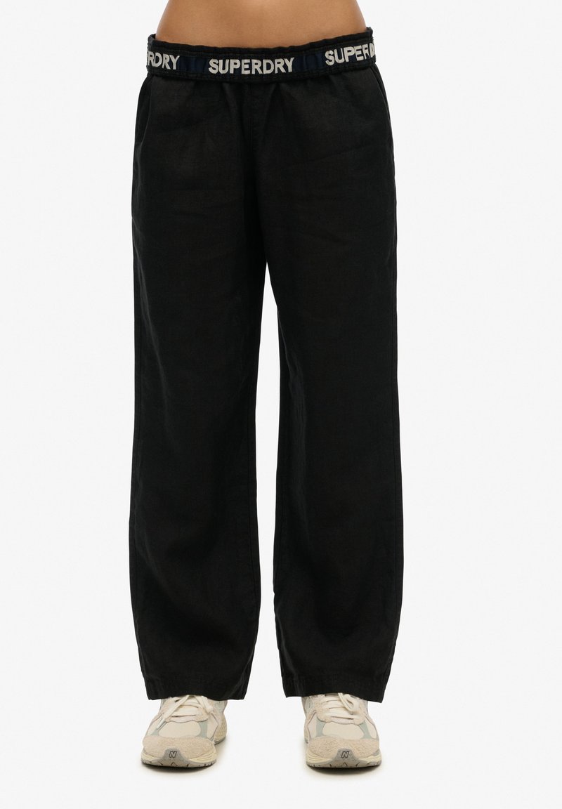 Pantalons larges en lin noir avec une taille élastique arborant le logo "SUPERDRY" en blanc et bleu. Tissu texturé et coupe décontractée.