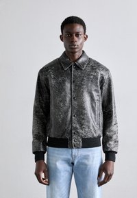 Sort, struktureret jakke med krave, trykknapper og ribstrik ved ærmerne. Båret over lyseblå jeans foran en neutral baggrund.