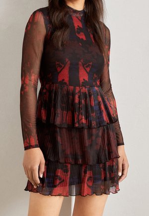 Femme portant une robe noire en mesh à manches longues avec des motifs abstraits d’arbres et de fleurs rouges et bleus, avec une jupe plissée à étages.