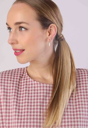 Un mannequin avec de longs cheveux blonds raides porte un haut en vichy rose et blanc. Elle a une boucle d'oreille dorée avec une petite barre blanche. Peau claire.