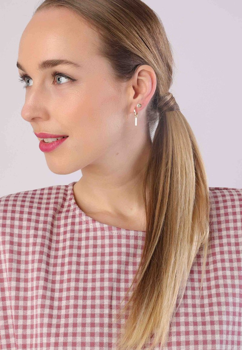 Model met lang, steil blond haar draagt een top in roze en witte ruitjes. Ze heeft een gouden ringoorbel met een kleine witte staaf. Heldere huid.