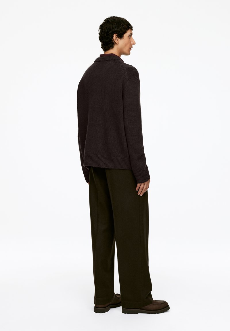 Dunkelbrauner Strickpullover mit hohem Kragen, lockerer Passform und gerippten Bündchen, kombiniert mit weit geschnittenen schwarzen Hosen und dunkelbraunen Schuhen.