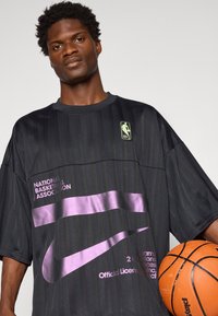 Black short-sleeve jersey with vertical stripes, featuring a purple Nike swoosh and NBA logo. Includes text detailing brand and licensing.