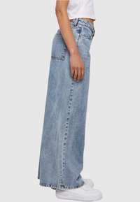 Falda maxi de denim azul claro con corte holgado, bolsillos laterales, dobladillo deshilachado y cintura alta. Combinada con zapatillas blancas.