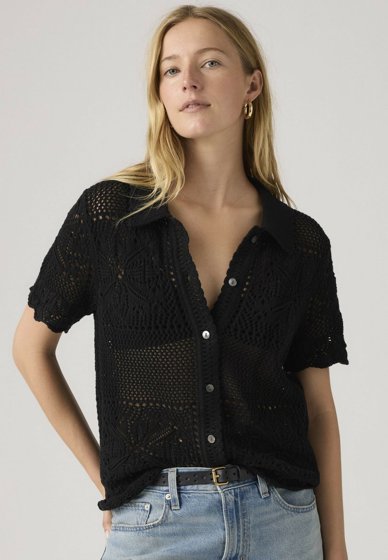 Blonde vrouw met gouden oorringen, een zwart gehaakt kortemouwig hemd met knopen, blauwe spijkerbroek en een zwarte riem met een gouden gesp.