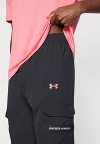 Μαύρα cargo σορτς με ελαστική μέση, με λογότυπο Under Armour σε κοραλί και πλαϊνή τσέπη. Μερικώς  ορατό ανοιχτό ροζ πουκάμισο.