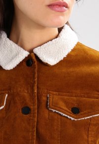 Bruine ribfluwelen jas met een witte sherpa-kraag, voorzien van zwarte knopen en twee voorzakken met witte stikselaccenten.