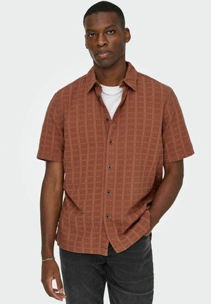Mann iført en rustfarget kortermet skjorte med subtilt rutemønster og svarte jeans, stående med en hånd i lomma.