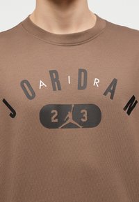 Καφέ μπλουζάκι με το κείμενο "JORDAN" σε μεγάλες γράμματα πάνω από μια μαύρη οβάλ επιφάνεια που περιέχει τους αριθμούς "2" και "3" με τη σιλουέτα ενός παίκτη μπάσκετ.