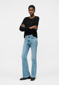 Woman stands with arms crossed wearing black sweater, light blue straight-leg jeans, and black shoes against plain background.