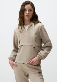 Felpa beige con cappuccio, caratterizzata da una zip frontale a mezza lunghezza, polsini elasticizzati e lacci decorativi. Tessuto morbido e testurizzato.