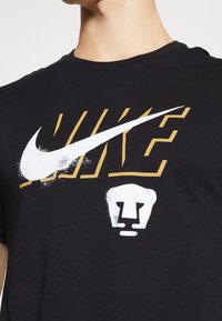 T-shirt en coton noir avec un grand logo Nike blanc et un texte "NIKE" doré, accentué par un graphique de lion blanc en dessous.