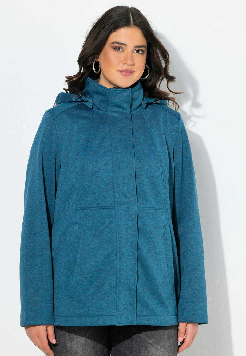 Nainen, joka käyttää teal-väristä takkia, jossa on korkea kaulus ja pitkät hihat, seisoo valkoista taustaa vasten.