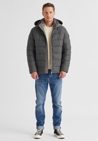 Graue Pufferjacke mit strukturiertem Oberteil, schwarzen Ärmeln und grauer Kapuze. Tragen über einem cremefarbenen Strickpullover und blauen Jeans.