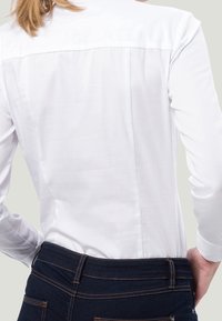 Camicia bianca a maniche lunghe con una texture liscia, dotata di una spalla sul retro, abbinata a jeans in denim scuro con cuciture visibili.