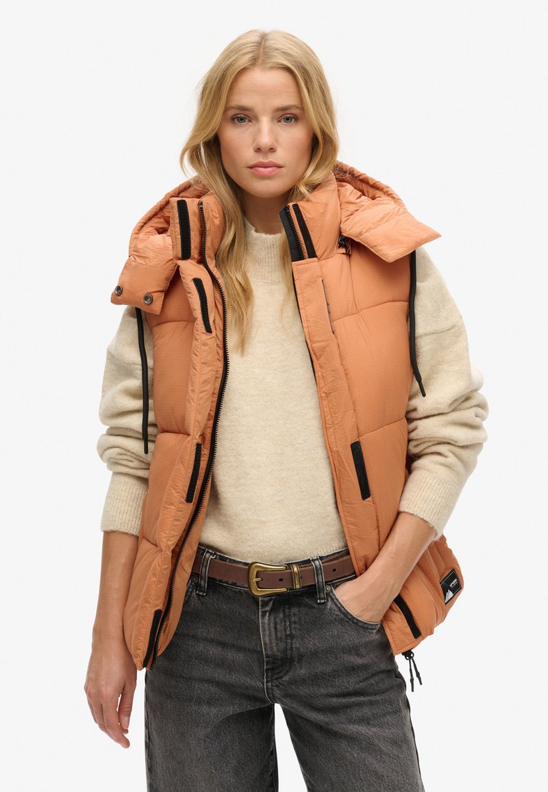 Blondynka w beżowym swetrze, pomarańczowej pikowanej kamizelce z czarnymi szczegółami, ciemnoszarych dżinsach i brązowym pasku, stoi na tle białego tła.