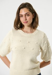 Cremefarvet striktrøje med korte, puffede ærmer. Har strukturerede boble detaljer og ribbet kant. Blødt, chunky garn og rund halsdesign.