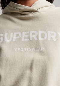 Béžová mikina s kapucí vyrobená z jemného bavlněného materiálu, s velkým bílým nápisem "SUPERDRY" a "SPORTSWEAR" napříč přední částí.