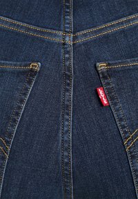 Mörkblå denimjeans med kontrasterande gula sömmar, utrustade med två bakfickor och en liten röd Levi's-etikett på den högra fickan.