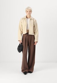 Beige zkrácená fleecová bunda s dvěma předními kapsami, spárovaná s hnědými kalhotami s širokými nohavicemi a držící malou černou kabelku. Hladká, texturovaná tkanina.