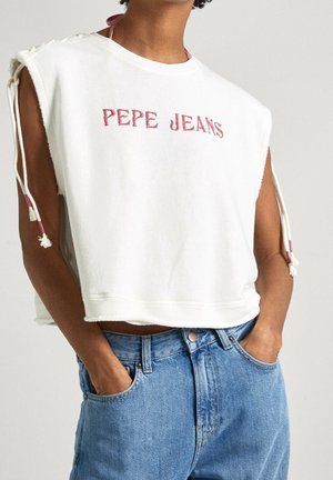 Person iført en hvid ærmeløs top med "PEPE JEANS" i pink tekst og blå cowboybukser, hænder i lommerne, neutral baggrund.