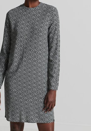 Kvinde iført en sort og hvid langærmet kjole med et lille blomsterprint, stående mod en ensfarvet lys grå baggrund.