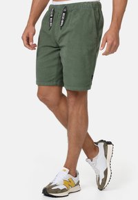 Kaki-grüne Cordshorts mit elastischem Bund, schwarzen Kordeln und Seitentaschen. Kombiniert mit weißen Sneakers mit gelben Akzenten.