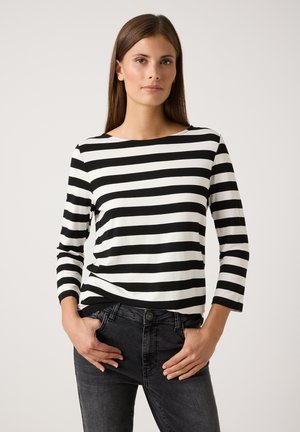 Woman wearing black and white horizontal striped long-sleeve shirt with black jeans, standing with hands near pockets against plain background.