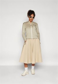 Görbén hajú nő napszemüveget visel, bézs zipzáros kabátot, széles bézs szoknyát, fehér zoknit és krém színű sportcipőt, háttérben egy sima felülettel.