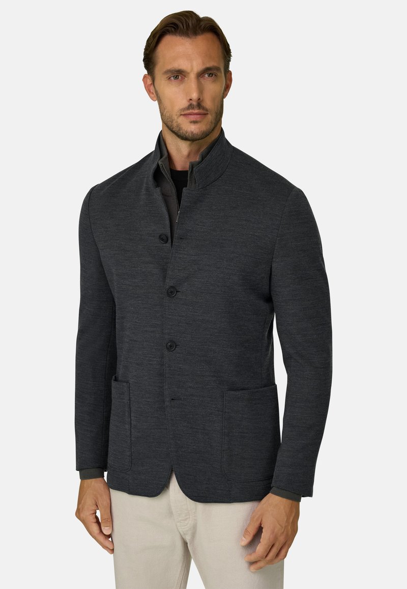 Homme portant une veste boutonnée gris foncé avec un col montant et des poches avant, associée à un pantalon beige clair.