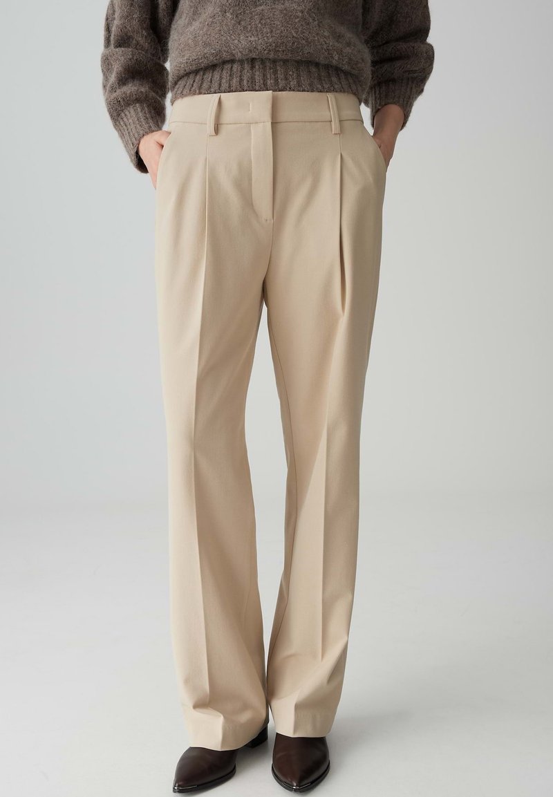 Beige, hoge taille broeken met plooien, gemaakt van een zachte stof. Bevatten een getailleerde pasvorm en zijzakken. Gecombineerd met bruine puntige schoenen.