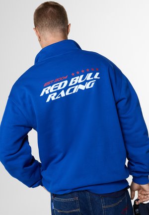 Persona que lleva una chaqueta azul con "Red Bull Racing EST. 2004" y estrellas rojas estampadas en la espalda, sosteniendo un objeto negro en la mano derecha.