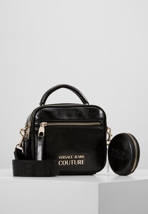 Sort lædertaske fra Versace Jeans Couture med gyldent lynlås, aftagelig rem, top-håndtag og lille rund møntlomme på hvid overflade.