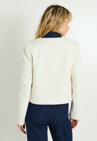 Chaqueta corta blanca texturizada con cuello redondo y mangas largas, combinada con pantalones de mezclilla oscuros. Diseñada con una silueta limpia y estructurada.