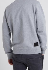 Man som bär en grå sweatshirt och mörka jeans med handen i fickan, sedd bakifrån mot en enfärgad bakgrund.