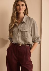 Light gray button-up shirt with two front pockets, rolled sleeves; paired with maroon pinstriped trousers and a brown belt.