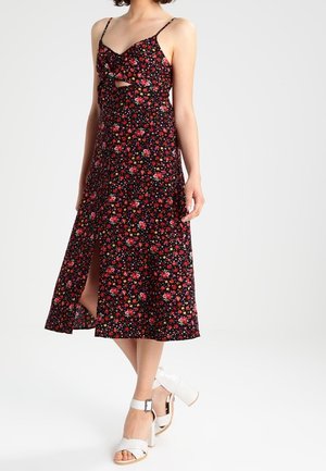 Abito midi floreale nero con spalline regolabili, dettaglio con nodo frontale e spacco laterale. Indossato con sandali bianchi con tacco a blocco.